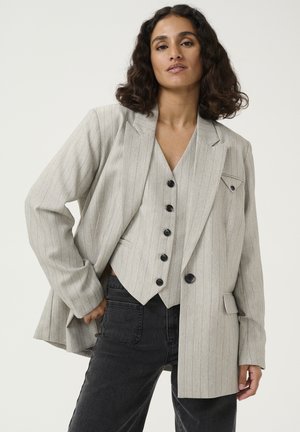 Svetle šedý blazer s jemným proužkem a detaily na kapsách, doplněný o odpovídající vestu s černými knoflíky, nošený přes černé džíny.