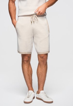 Mann trägt beige gestrickte Shorts mit Kordelzug und weiße Sneaker, steht mit den Händen in den Taschen vor einem einfarbigen Hintergrund.