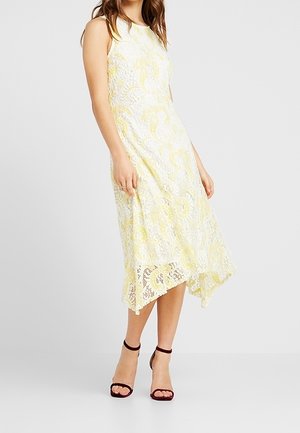 Spitzen-Midikleid in Gelb mit filigranen Blumenmustern, mit ärmellosem Design und asymmetrischem Saum. Kombiniert mit Riemchen-Pumps in Burgunderrot.