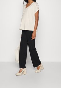 Jasnobeżowa bluzka z dekoltem w serek i koronkowymi akcentami na rękawach, w zestawie z czarnymi, szerokimi spodniami i kremowymi butami na masywnym obcasie, trzymająca beżową torbę typu tote.