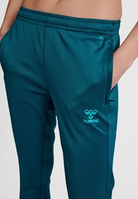 Turquoise sportbroeken met een gladde textuur, voorzien van een tailleband en zijzakken. Het Hummel-logo is in contrasterend lichtblauw.
