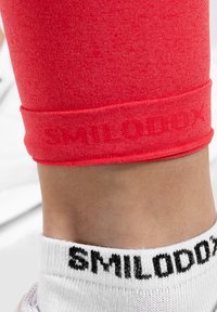 Rote sportliche Leggings mit glatter Textur, ausgestattet mit einem gedruckten Logo "SMILODAX" in passendem Farbton am Saum, kombiniert mit weißen Socken.