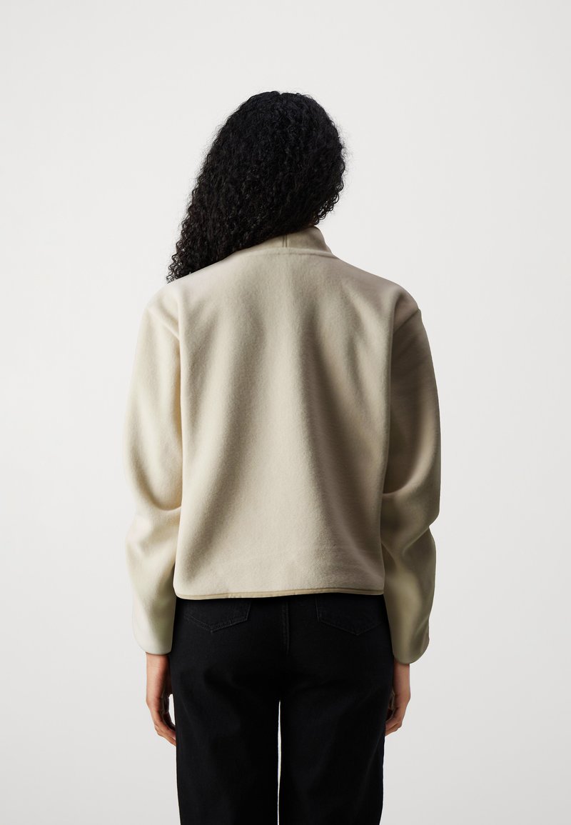 Persoon met krullend zwart haar, gekleed in een crèmekleurige fleece jack en zwarte broek, met de rug naar de camera tegen een effen witte achtergrond.