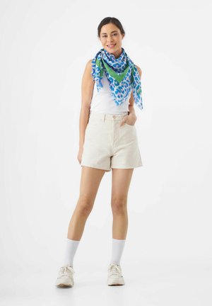 Beige katoenen shorts met zijzakken, gecombineerd met een kleurrijke driehoekige sjaal met een blauw en groen luipaardmotief, een wit tanktop en sneakers.