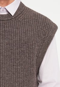 Gilet en tricot brun avec texture côtelée, design à larges épaules et col rond, superposé à une chemise blanche à col.