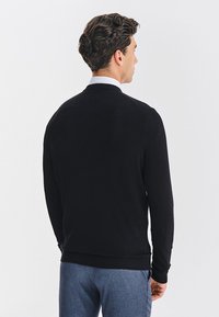 Czarny sweter z dzianiny, z okrągłym dekoltem i długimi rękawami; dopasowany krój z prążkowanym dołem i mankietami, gładka faktura; noszony na szarych spodniach.