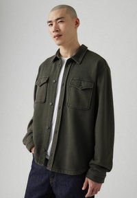 Camisa verde abotonada con cuello, bolsillos delanteros y mangas largas. Hecha de tela suave, con un ajuste relajado y detalles de costuras visibles.