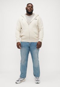 Licht beige zip-up hoodie met een zachte textuur, gecombineerd met lichtblauwe jeans. Sneakers hebben grijze accenten en een chunky ontwerp.