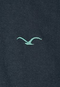 Dunkelblauer Pullover mit weicher Textur, versehen mit einem hellgrünen, gestickten Möwenlogo auf der Vorderseite. Einfaches, rundes Ausschnitt-Design.