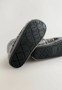 Grijze wollen slippers met een textured stoffen bovenkant, voorzien van een rubberen loopzool met geometrische patronen. Zachte voering zichtbaar bij de opening.