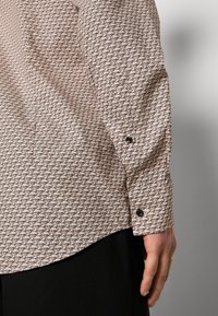 Camicia beige chiaro con motivo di testo nero ripetuto. Presenta polsini con bottoni e un orlo curvo, realizzata in un tessuto liscio. Pantaloni neri visibili sotto.
