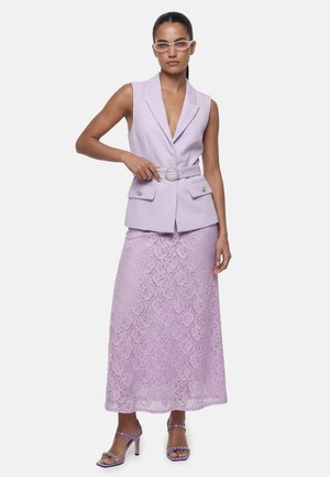 Kobieta ubrana w bezrękawowy liliowy blezer z dekoracyjnym paskiem, pasującą koronkową maxi spódnicę, liliowe sandały z ozdobami i liliowe okulary.