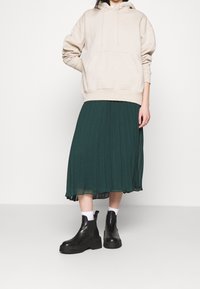 Beige hoodie med en framficka, parat med en grön plisserad kjol och svarta plattform ankelstövlar. Outfiten har kontrasterande texturer.