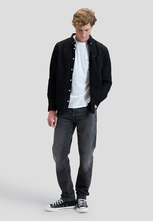 Jeune homme portant une chemise noire boutonnée sur un t-shirt blanc, un jean gris et des baskets noires, debout, une main dans la poche, regardant vers le bas.