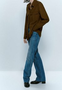 Brown long-sleeve shirt with two chest pockets, paired with light blue flared jeans and black ankle boots. Plain background.