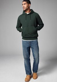 Dark green hooded sweatshirt with a front pouch pocket, paired with blue denim jeans and brown shoes, featuring a relaxed fit and drawstrings.