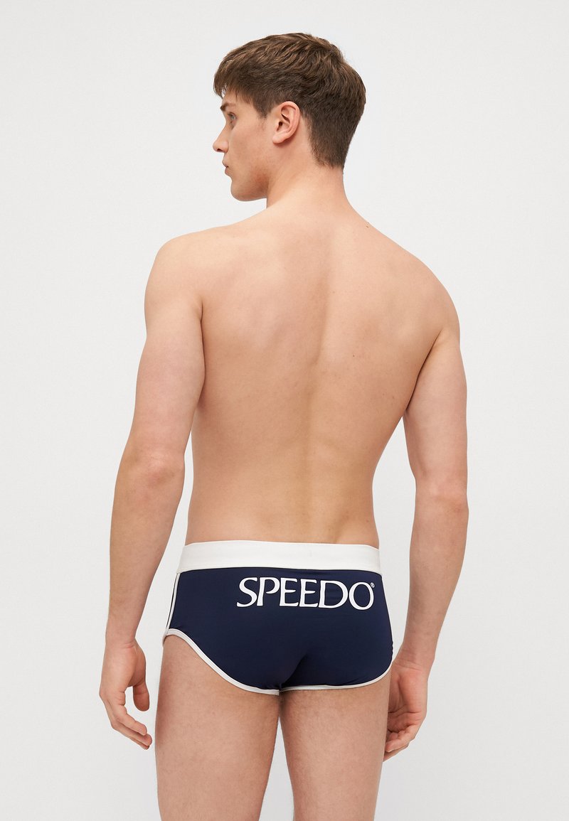 Sötétkék úszónadrág nylonból, fehér elasztikus derékpánttal. A hátulján nagy fehér betűkkel szerepel a "SPEEDO" logó.