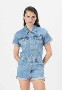 Combinaison en denim à manches courtes et poignets retroussés. Lavage bleu clair, avec fermeture à boutons et poches avant pour le détail.