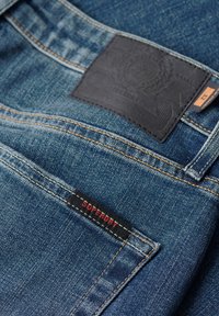 Vaqueros de mezclilla en azul oscuro con costuras naranjas en contraste, presentando un parche de cuero negro con grabado y una etiqueta roja "SUPERDRY".