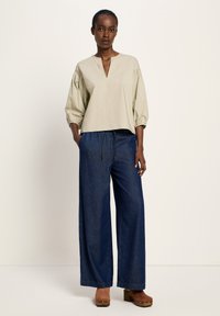 Frau steht in einer beigen lockeren Bluse mit 3/4-Ärmeln, weiten dunklen Jeans und braunen Plateauschuhen.