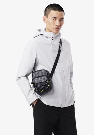 Joven vestido con una chaqueta con capucha de color gris claro, pantalones negros y un bolso de bandolera negro con patrones geométricos y acentos amarillos.