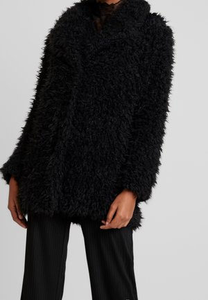 Person iført en sort, fluffy tekstureret frakke over sorte bukser, stående mod en ensfarvet grå baggrund.