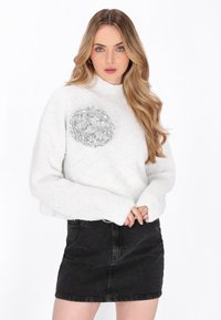 Witte, chunky gebreide trui met een hoge hals en een zilveren pailletten bloemaccent op de borst, gecombineerd met een zwarte denim rok.