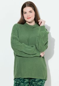 Maglione lavorato a maglia verde con motivi testurizzati sulle maniche e un collo alto. Abbinato a pantaloni verdi con motivo fogliare su uno sfondo neutro.