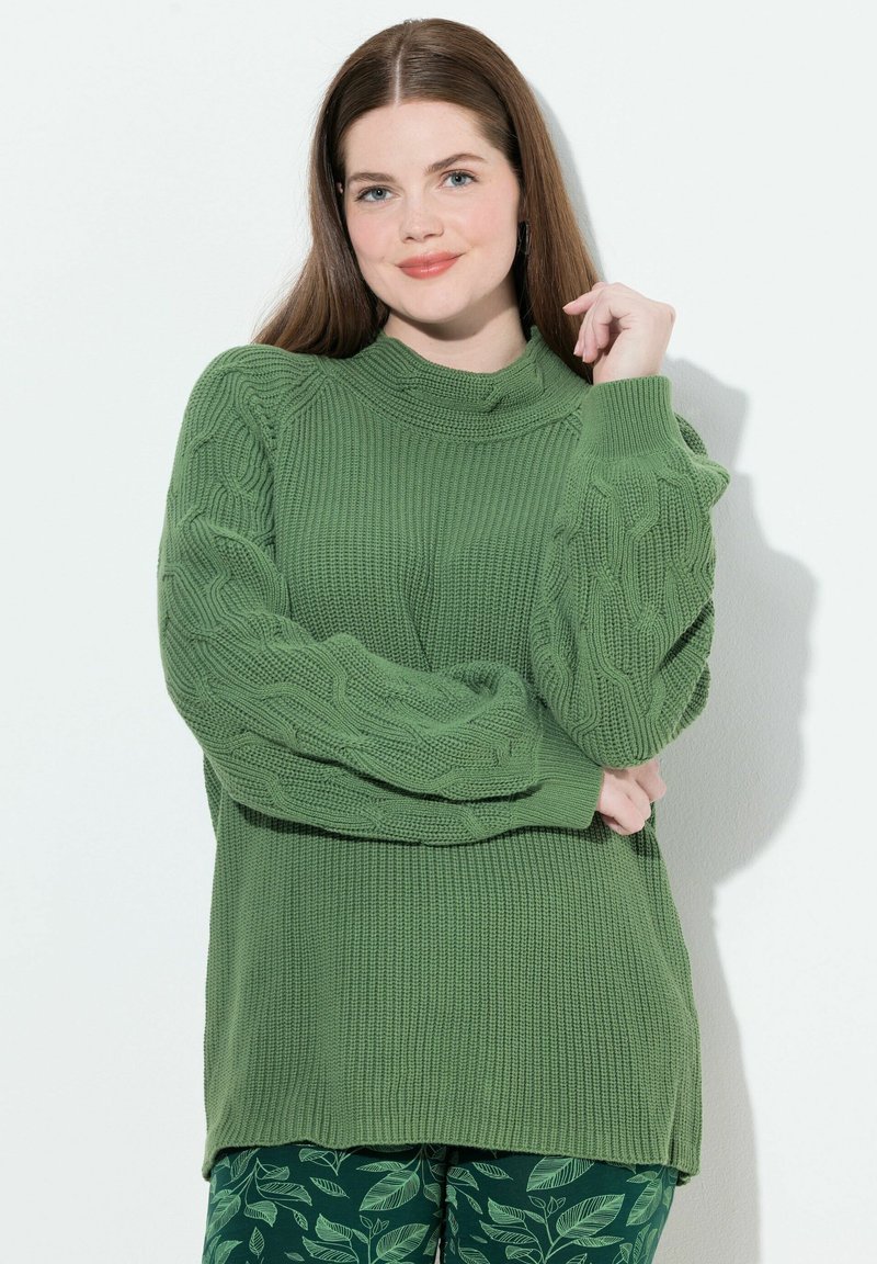 Maglione lavorato a maglia verde con motivi testurizzati sulle maniche e un collo alto. Abbinato a pantaloni verdi con motivo fogliare su uno sfondo neutro.