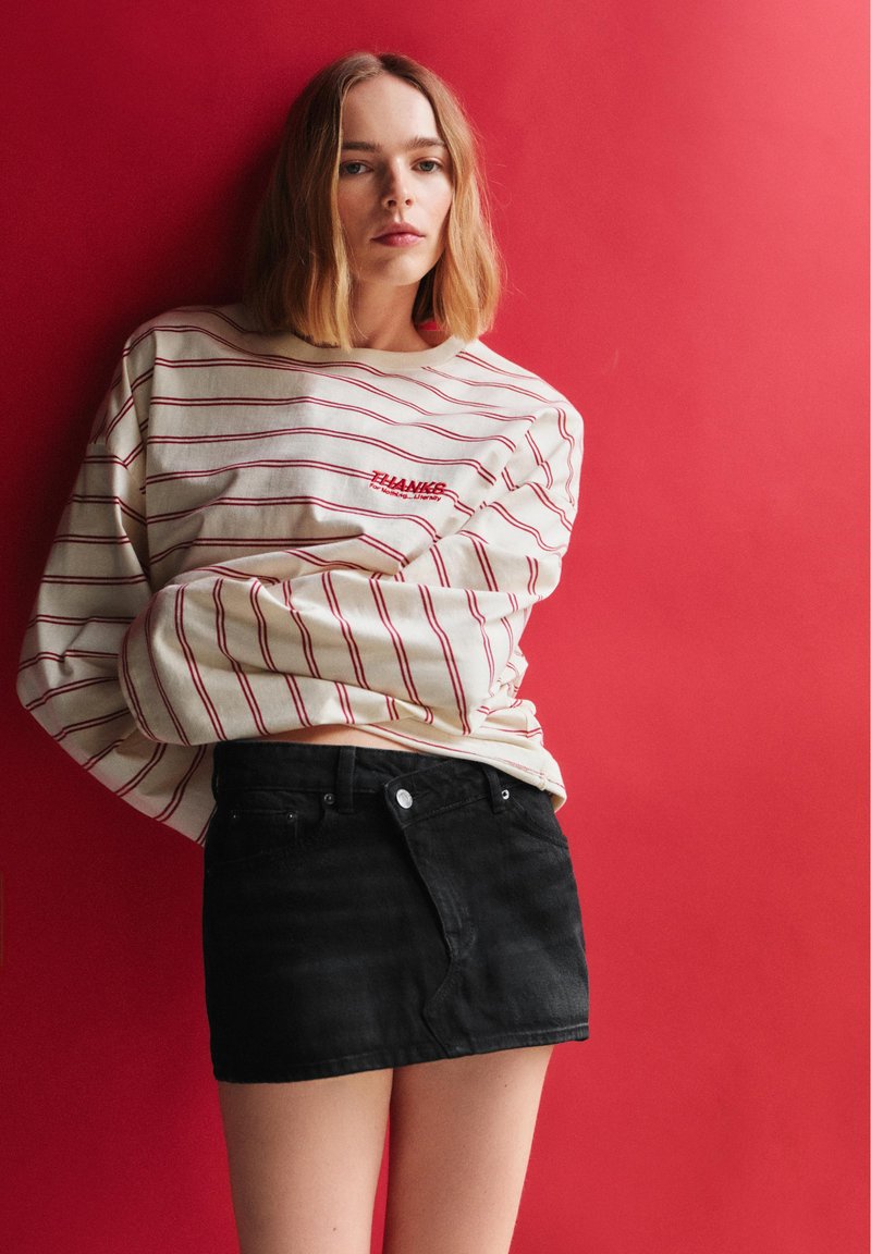 Jeune femme portant un haut rayé à manches longues et une mini-jupe noire, debout contre un mur rouge avec une expression neutre.