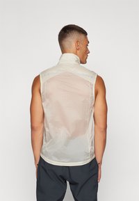 Bezrukávový svetlobežový vest s vysokým golierom, vyrobený z priesvitného materiálu s ventilačnými otvormi na chrbte a priliehavým strihom.