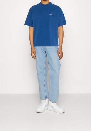 Mand iført en blå T-shirt med korte ærmer og lille hvidt logo, lyseblå jeans og hvide høje sneakers, stående mod en hvid baggrund.