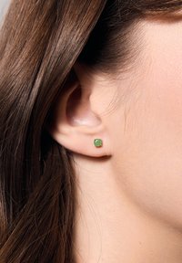 Małe zielone kamienie szlachetne wkręty osadzone w metalowym stawie, umieszczone w płatku ucha w zestawieniu z prostymi, brązowymi włosami. Minimalistyczny design i okrągły kształt.