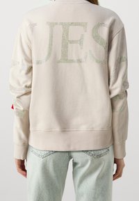 Vaalea beige collegepaita, jossa on takana suuret strassikirjaimet. Pitkissä hihoissa kimaltelevat yksityiskohdat. Resorit helmassa ja hihojen manseteissa. Vaaleat farkut.