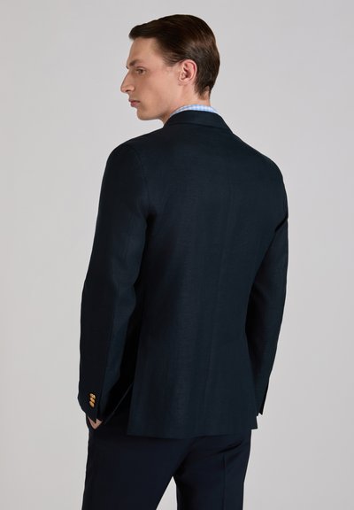 Mann, der nach links schaut, trägt eine dunkelblaue Anzugjacke mit goldenen Knöpfen und ein hellblaues kariertes Hemd vor einem einfarbigen grauen Hintergrund.