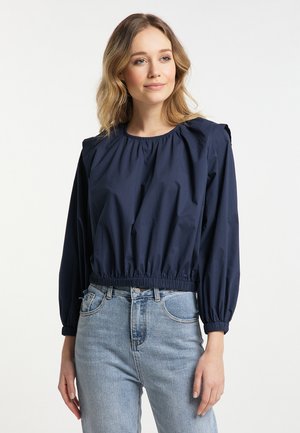Junge Frau mit welligem blondem Haar trägt eine dunkelblaue, langärmelige, geraffte Bluse und hellblaue, hoch taillierte Jeans vor weißem Hintergrund.