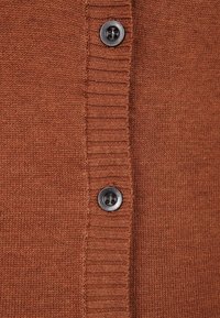 Rostfärgad stickad tyg med ribbad knapplist och två mörkbruna fyrhålsknappar knäppta.