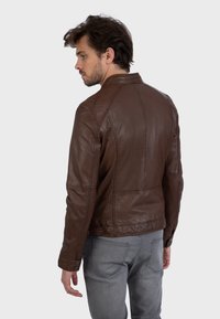 Brown leather jacket with a fitted design, featuring a stand collar, seam detailing, and button accents. Worn over grey pants.
