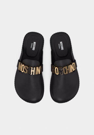 Sandales noires sans lacets avec des lettres métalliques dorées formant le mot "Moschino" sur les brides, semelles rembourrées, et "Moschino Milano" imprimé à l'intérieur.