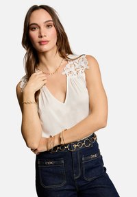 Top sin mangas de color blanco con detalles de encaje en los hombros, diseño de escote en V, combinado con jeans de mezclilla oscura y accesorios en tono dorado, incluyendo pulseras y un cinturón.