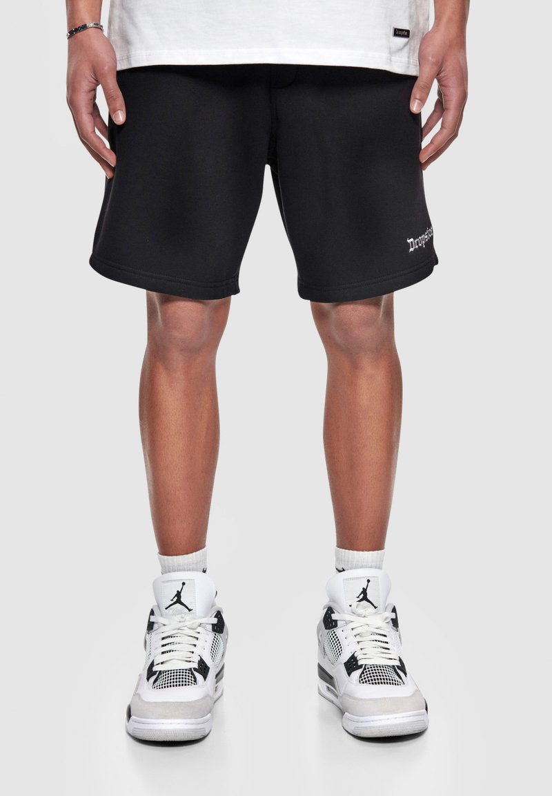 Schwarze Shorts mit glatter Textur, versehen mit einem kleinen weißen Textlogo. Kombiniert mit weißen und schwarzen Sportschuhen mit Netzeinsätzen.
