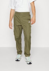 Oliivinvihreät cargo-housut, joissa on joustava vyötärönauha, useita taskuja ja valkoinen Nike-logo vasemmassa reisiosassa. Yhdistetty vaalean aqua-sävyisiin tennareihin.