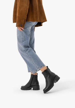 Personne portant un jean bleu, une veste marron et des bottines noires à talons carrés, debout avec un pied légèrement levé.