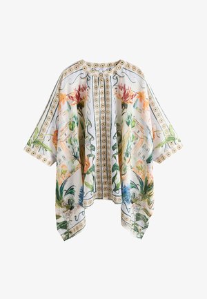 Tânără cu părul ud purtând o vestă florală și chiloți de bikini maro stă pe o plajă cu nisip, cu oceanul în fundal.