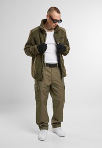 Chaqueta de forro polar de color verde oliva con un bolsillo con cremallera, combinada con pantalones cargo beige que cuentan con múltiples bolsillos, y zapatillas blancas.