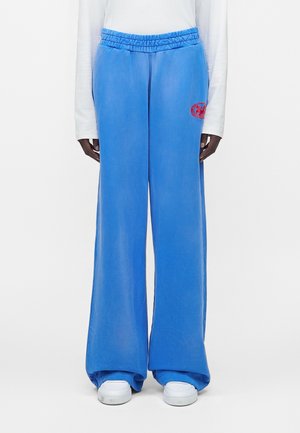 Osoba ubrana w jasnoniebieskie spodnie o szerokich nogawkach z czerwonym okrągłym logo na lewym udzie, białą koszulę z długim rękawem i białe trampki.