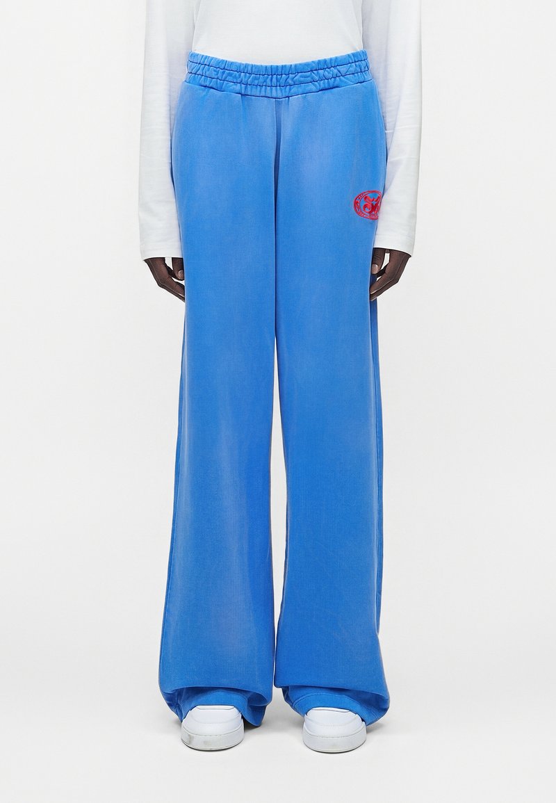 Άτομο με φωτεινό μπλε παντελόνι φαρδιάς γραμμής με κόκκινο στρογγυλό λογότυπο στο αριστερό μηρό, λευκό μακρυμάνικο πουκάμισο και λευκά αθλητικά παπούτσια.