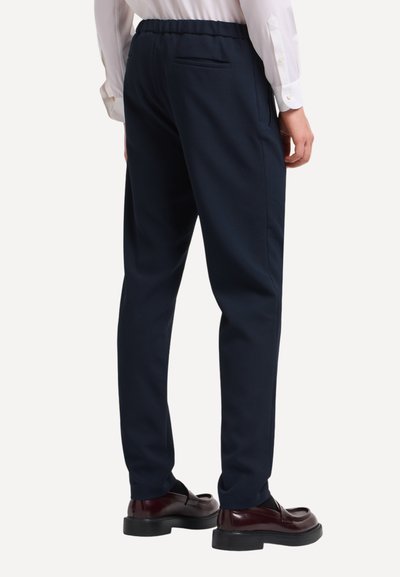Uomo vestito con pantaloni su misura di colore blu navy, camicia bianca e mocassini in pelle marrone scuro, in piedi con la schiena rivolta verso l'osservatore.