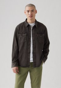 Cămașă din denim gri închis cu guler ascuțit, două buzunare la piept și închidere cu nasturi, purtată peste o cămașă albă și asociată cu pantaloni verzi.