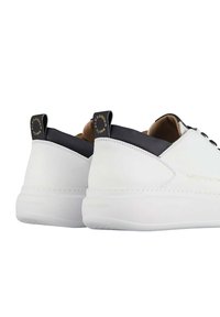 Par hvid lavtop-sneakers med sorte hælflapper og guld "Alexander Smith" branding på hæl og side, vist fra bagvinkel.